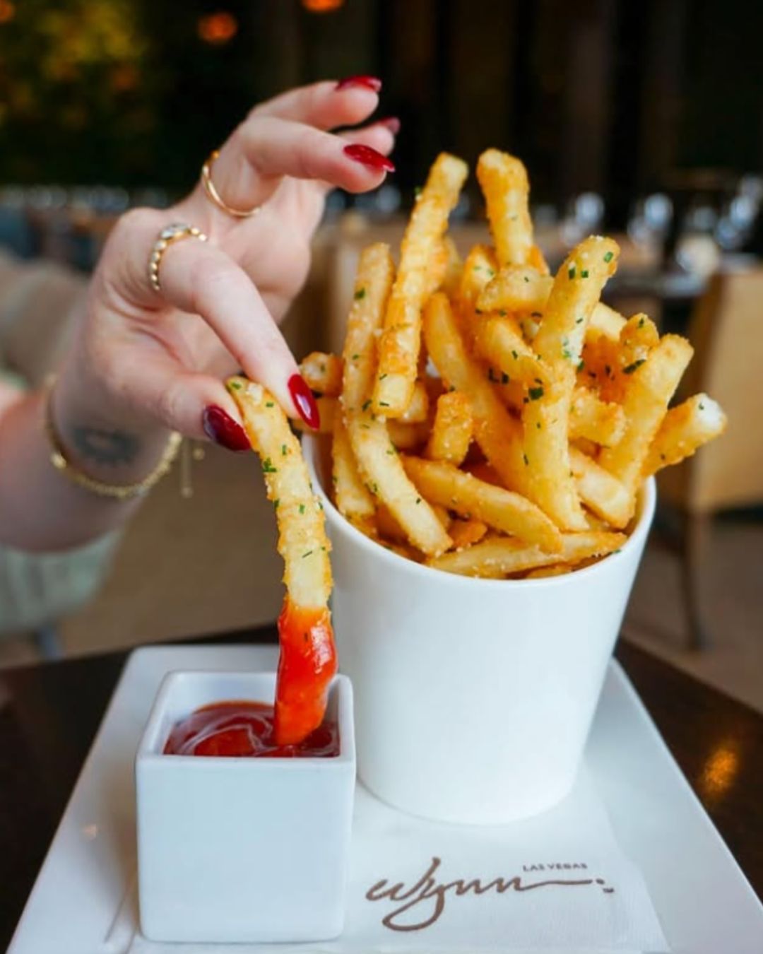 A server in a white butler jacket offers a plate of mini short rib hash with a quail egg to a smiling woman on a sun-drenched, European-style patio at La Cave, Wynn Las Vegas.